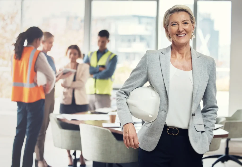 Confident woman in business attire knows her business debtor finance is under control, she holds a white hard hat, standing in an office with a group of people in safety vests and business clothes talking in the background