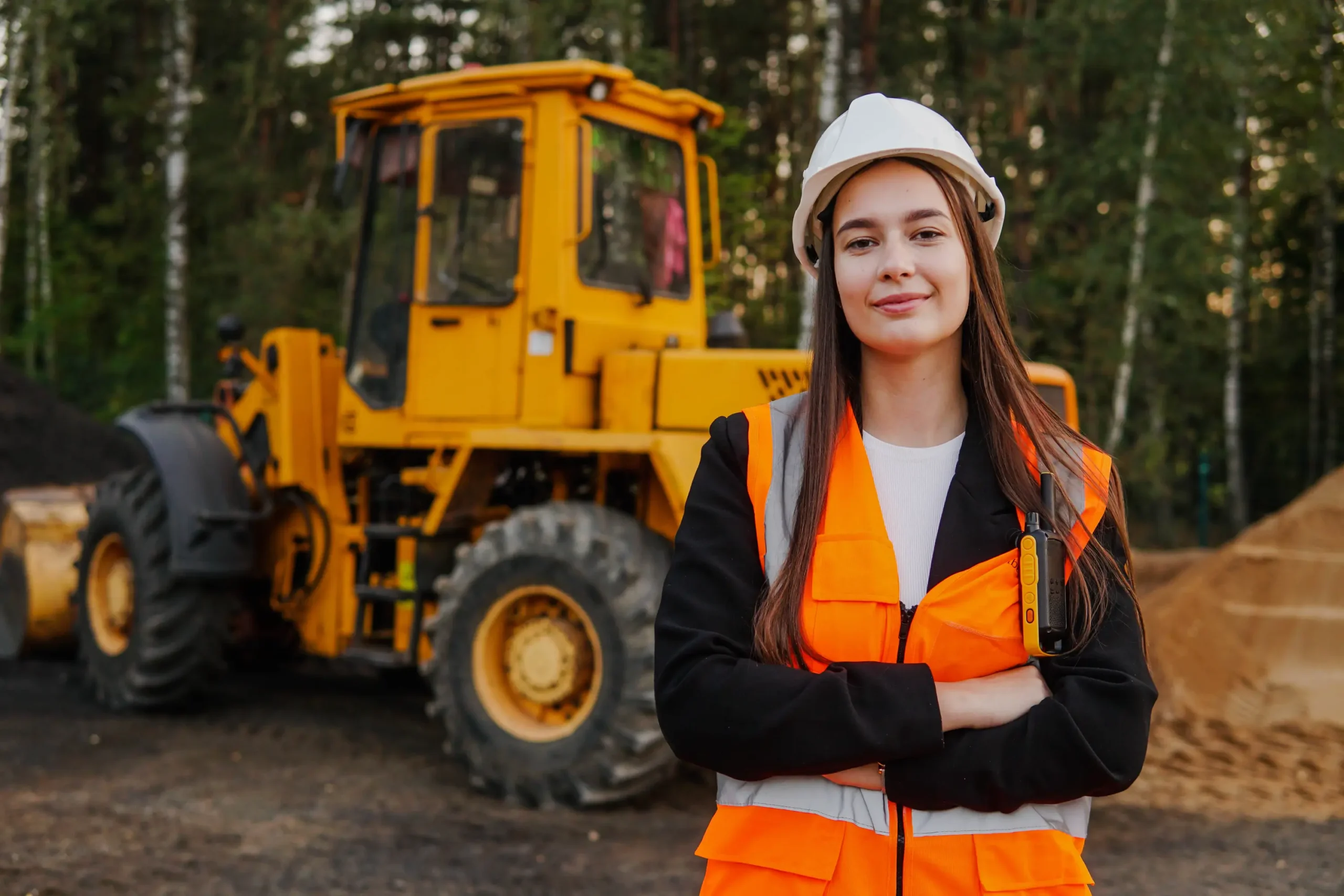 A woman wearing a white hard hat and orange safety vest stands with arms crossed in front of a yellow construction vehicle that was purchased with Plant machinery Finance