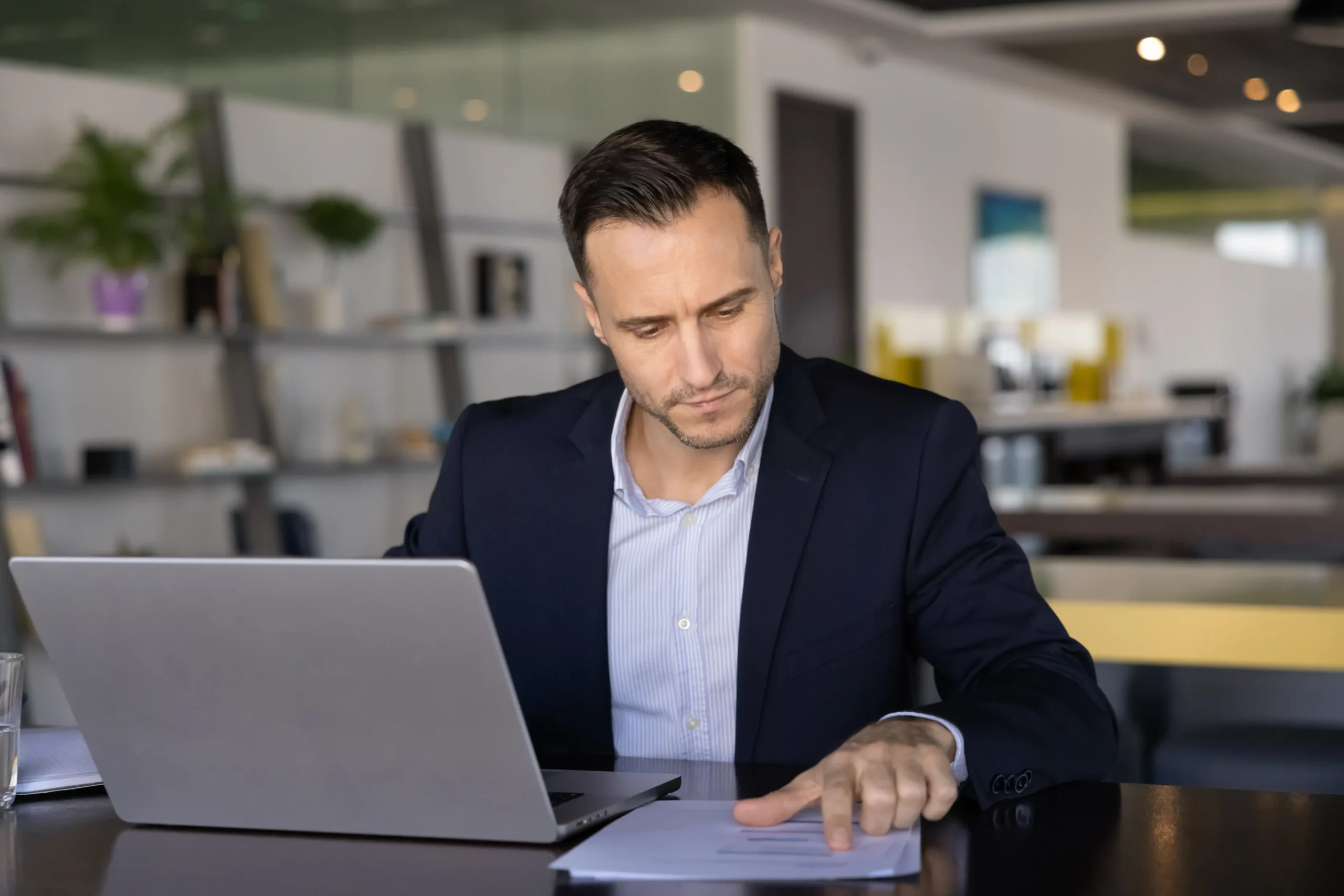 A man in a suit sits at a desk with a laptop, reading a document about an Unsecured business loans