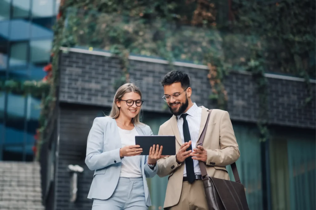 Two business professionals stand outdoors, smiling as they look at a tablet with modern buildings and greenery behind them. Contact Borrow Bridge to learn more.