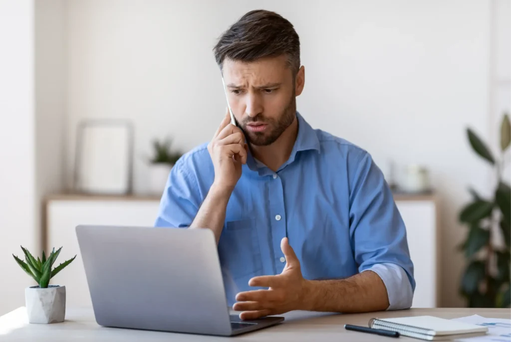A man in a blue shirt sits at a desk with a laptop, talking on the phone and gesturing with concern, as he discusses making bad credit personal loans easy.