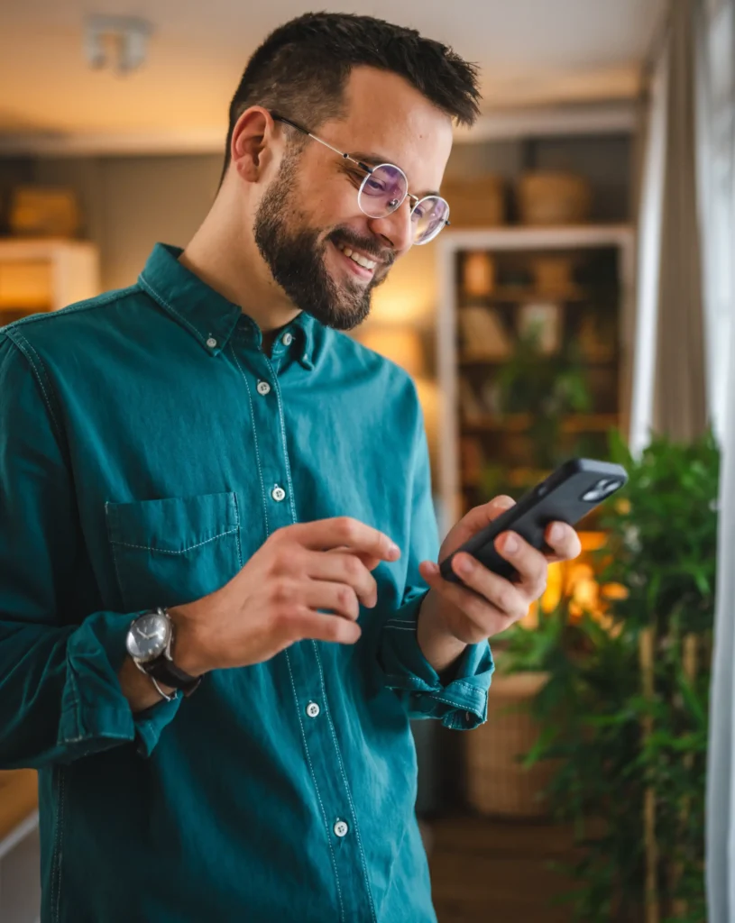 A man wearing glasses and a teal shirt stands indoors, smiling whilst looking at his mobile phone, checking his Personal Cash Loan details.