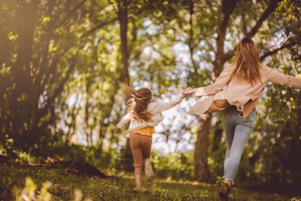 An adult and a child are holding hands and running through a sunlit, green forest with trees and grass, enjoying carefree moments funded by a personal loan for education expenses.