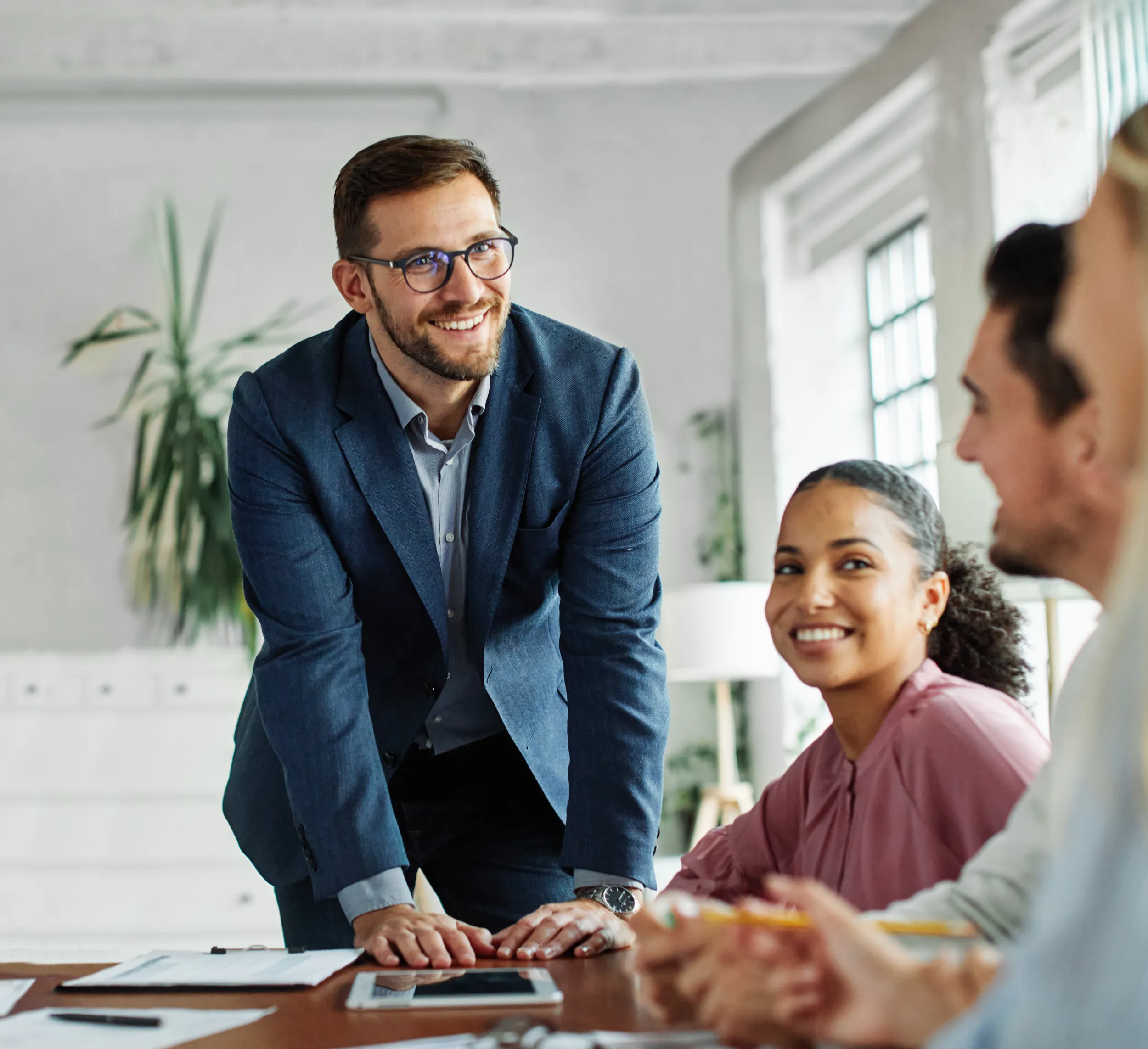 A man in a blue suit stands and leans on a table, smiling at three seated colleagues during a meeting in a bright Brisbane office, discussing personal loan options with Borrow Bridge.