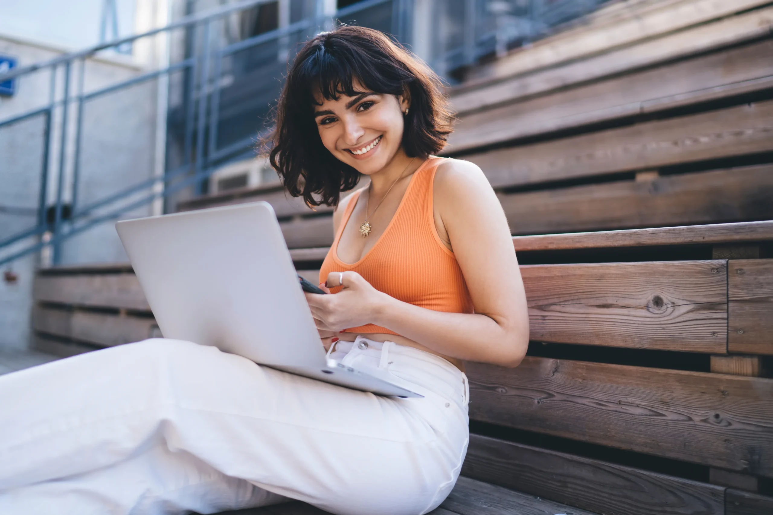 A woman in an orange vest top and white trousers sits on outdoor wooden steps in Brisbane, smiling at the camera while using a laptop and holding a mobile, exploring Wedding Loan Finance options with Borrow Bridge.